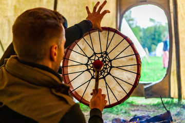 Wall Mural - Fusion of cultural & modern music event. An over the shoulder view of a spiritual man playing native drum as he sits inside a teepee tent during a celebration of Native American culture, room for copy