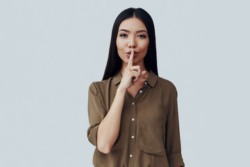 Keep my secret. Beautiful young Asian woman keeping finger on lips while standing against grey background