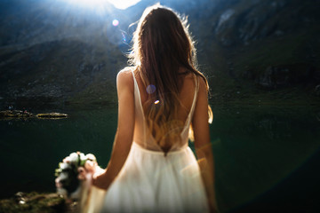 Wall Mural - Unrecognizable back view of a young long haired bride walking against sunrise in mountains holding a bouquet of flowers.