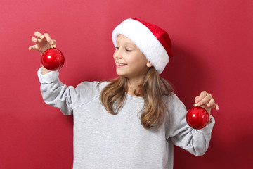 Wall Mural - Cute girl on a colored background in a Christmas hat holds balls to decorate the Christmas tree