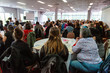 © Valmedia - Atmosphere during corporate conference. A rear view of three female co-workers sitting at a round table during a training event for a large-scale business. Blurry colleagues are seen in the background