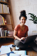 Young beautiful smiling woman with dark curly hair sitting on floor with laptop on knees happily looking aside with books and cup of coffee to go near at home