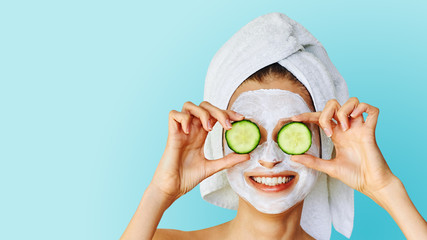 Beautiful young woman with facial mask on her face holding slices of cucumber. Skin care and treatment, spa, natural beauty and cosmetology concept.