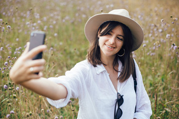 Wall Mural - Hipster girl with backpack taking selfie on phone while relaxing in wildflower meadow, traveling in sunny mountains. Stylish woman in hat enjoying hiking on top of mountain. Summer vacation