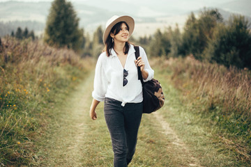 Wall Mural - Happy hipster girl with backpack traveling on top of sunny mountain, walking on hills with woods. Stylish woman smiling and  enjoying hiking in mountains. Wanderlust and travel.