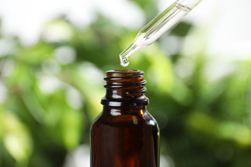 Dropper with essential oil over bottle against blurred background, closeup