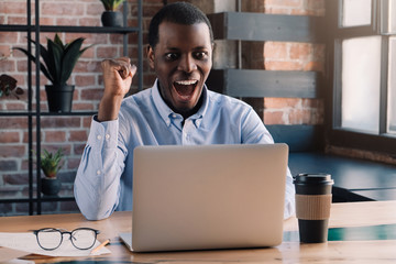 Young african american shocked business man using laptop computer, making winner gesture with fist