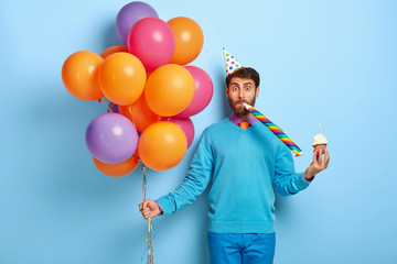 Funny man with bristle, wears party hat, blows horn, holds sweet muffin with burning candle, holds bunch of multicolored balloons, going to have birthday celebration, isolated on blue background