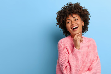 Charismatic beautiful young Afro woman touches chin gently, smiles broadly and shows white teeth, looks aside, enjoys friendly company, wears loose rosy sweater, models against blue background.