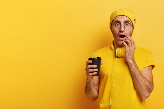 Image of emotional youngster holds disposable cup of coffee, stares with widely opened eyes, cannot believe eyes, wears yellow hat, t shirt, reacts on shocking news, poses indoor, free space on left