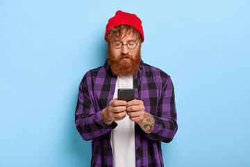 Fashionable hipster with red hair and thick beard, focused in smart phone, recieves link to some publication, wears red hat, purple checkered shirt, has leisure day, isolated on blue studio wall