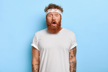 Indoor shot of surprised bearded man with red hair, stares with astonishment, has gymnastic every day, dressed in white casual clothes, headband, models over blue wall. People, emotions concept