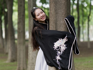 Beautiful Asian woman in black and white Chinese costume clothes hanfu hug tree trunk and smiling at camera in forest, traditional ancient Chinese beauty, time travel fiction.