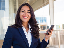 Happy Successful Business Professional Using Cellphone Outside. Beautiful Young Latin Woman In Office Suit Holding Smartphone And Smiling At Camera. Mobile Internet Concept