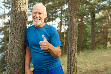 Cheerful attractive retired man with bald head and gray beard posing outdoors in sports clothes smiling happily, showing thumbs up gesture, choosing active healthy lifestyle, full of energy