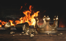 Golden Crown And A Goblet Full Of Gold On The King Table Over Burning Fire Background.