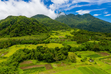 Wall Mural - Aerial drone view of clouds passing over lush greenfarmland with mountains and volcanos in the background (Camiguin, Philippines)