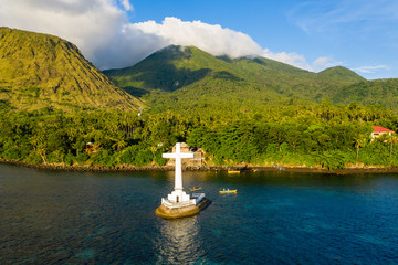 Wall Mural - Aerial view of golden evening sunlight illuminating a large cross marking a sunken cemetery off the volcanic tropical island of Camiguin