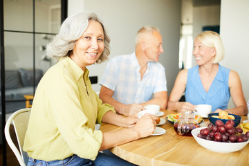 Wall Mural - Portrait of elegant mature woman sitting at dinner table and smiling at camera while enjoying lunch with friends and family, copy space