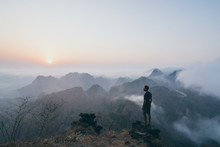 Man Standing On The Rock Overlooking Tropical Mountains At Sunrise Foggy Morning In Hpa-an, Myanmar