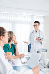 Young Vietnamese general practitionaer with tablet computer in hands discussing new innovative way of treatment with coworkers