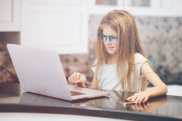 happy small smiling girl in glasses sitting in kitchen with laptop and cell phone online