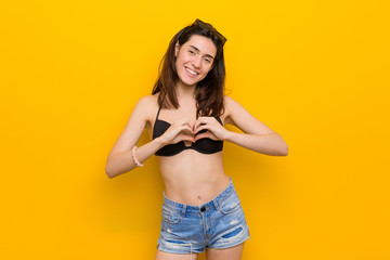 Wall Mural - Young brunette woman wearing a bikini against yellow background smiling and showing a heart shape with hands.