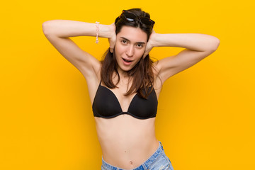 Wall Mural - Young brunette woman wearing a bikini against yellow background covering ears with hands trying not to hear too loud sound.