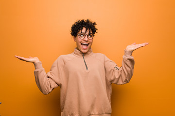 Young mixed african american woman against a brown background makes scale with arms, feels happy and confident.