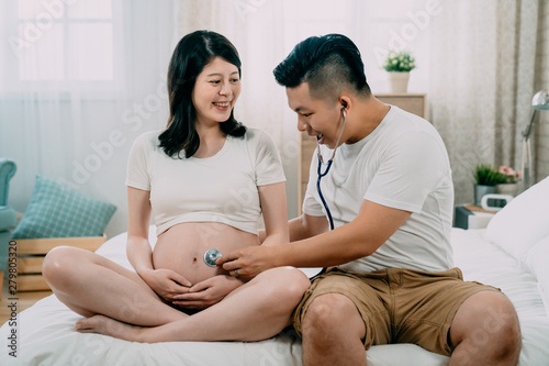 Young Pregnant Asian Japanese Woman And Handsome Husband Sitting On Bed Man Future Dad Using Stethoscope Listen To Wife Bare Belly With Unborn Baby Inside Happy Fathers Day Best Gift Ever