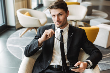 Photo of smart young man sitting in hotel hall with smartphone and eyeglasses during business trip