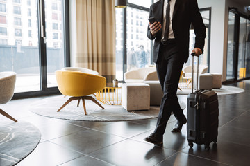 cropped photo of successful businessman wearing suit holding smartphone and walking with suitcase in