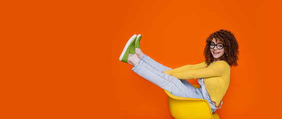 Cute curly haired hipster woman wearing in green shoes sitting on chair with funny raised legs