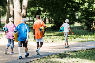 back view of happy senior and multicultural pensioners holding fitness mats and walking on grass