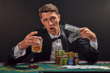 Handsome emotional man is playing poker sitting at the table in casino against a white spotlight.