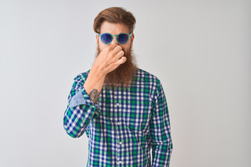 Wall Mural - Young redhead irish man wearing casual shirt and sunglasses over isolated white background smelling something stinky and disgusting, intolerable smell, holding breath with fingers on nose. Bad smells 