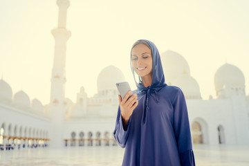 Wall Mural - Traveling by Unated Arabic Emirates. Pretty young woman in traditional abaya using smartphone in the Sheikh Zayed Grand Mosque, famous Abu Dhabi sightseeing.