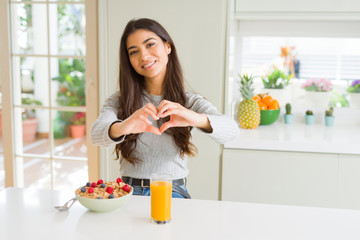 Wall Mural - Young woman eating healthy breakfast in the morning smiling in love showing heart symbol and shape with hands. Romantic concept.