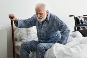 Wall Mural - Sad senior man with Alzheimer's disease holding walking stick and sitting on his bed in nursing home