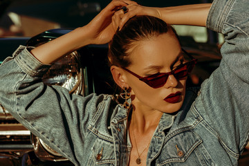 Outdoor close up fashion portrait of young beautiful fashionable woman wearing red cat eye sunglasses, denim jacket, leopard print earrings, necklace, posing near retro car, at the sunset