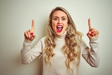 Beautiful woman wearing winter turtleneck sweater over isolated white background smiling amazed and surprised and pointing up with fingers and raised arms.