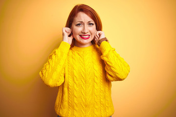 Sticker - Beautiful redhead woman wearing winter sweater standing over isolated yellow background covering ears with fingers with annoyed expression for the noise of loud music. Deaf concept.