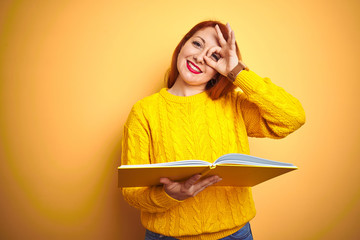 Poster - Young redhead student woman reading book standing over yellow isolated background with happy face smiling doing ok sign with hand on eye looking through fingers