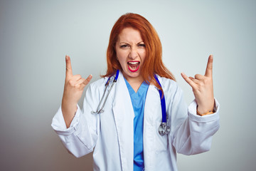 Sticker - Young redhead doctor woman using stethoscope over white isolated background shouting with crazy expression doing rock symbol with hands up. Music star. Heavy music concept.