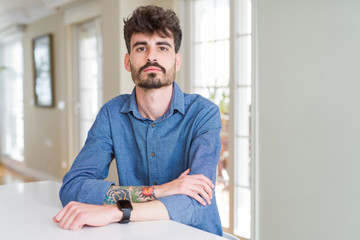 Sticker - Young man wearing casual shirt sitting on white table with serious expression on face. Simple and natural looking at the camera.