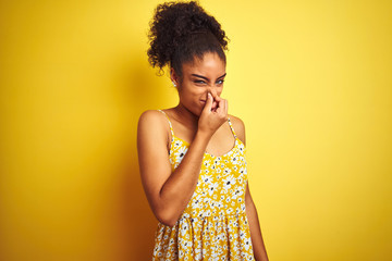 Sticker - African american woman wearing casual floral dress standing over isolated yellow background smelling something stinky and disgusting, intolerable smell, holding breath with fingers on nose. Bad smells