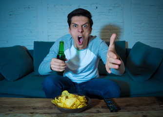 Wall Mural - Portrait of excited young man on couch watching football at night drinking beer. Sports fans concept