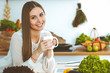 © rogerphoto - Young happy woman is holding white cup and looking at the camera while sitting at wooden table in the kitchen among green vegetables