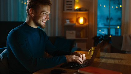 Evening at Home: Portrait of Handsome Man Sitting at His Desk Working on a Laptop. Smiling Freelancer Working on Computer. Side View Shot.