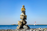 Fototapeta  - Tower of stones on the sea beach against the background of the lighthouse and the breakwater. Peace and tranquility in the open air